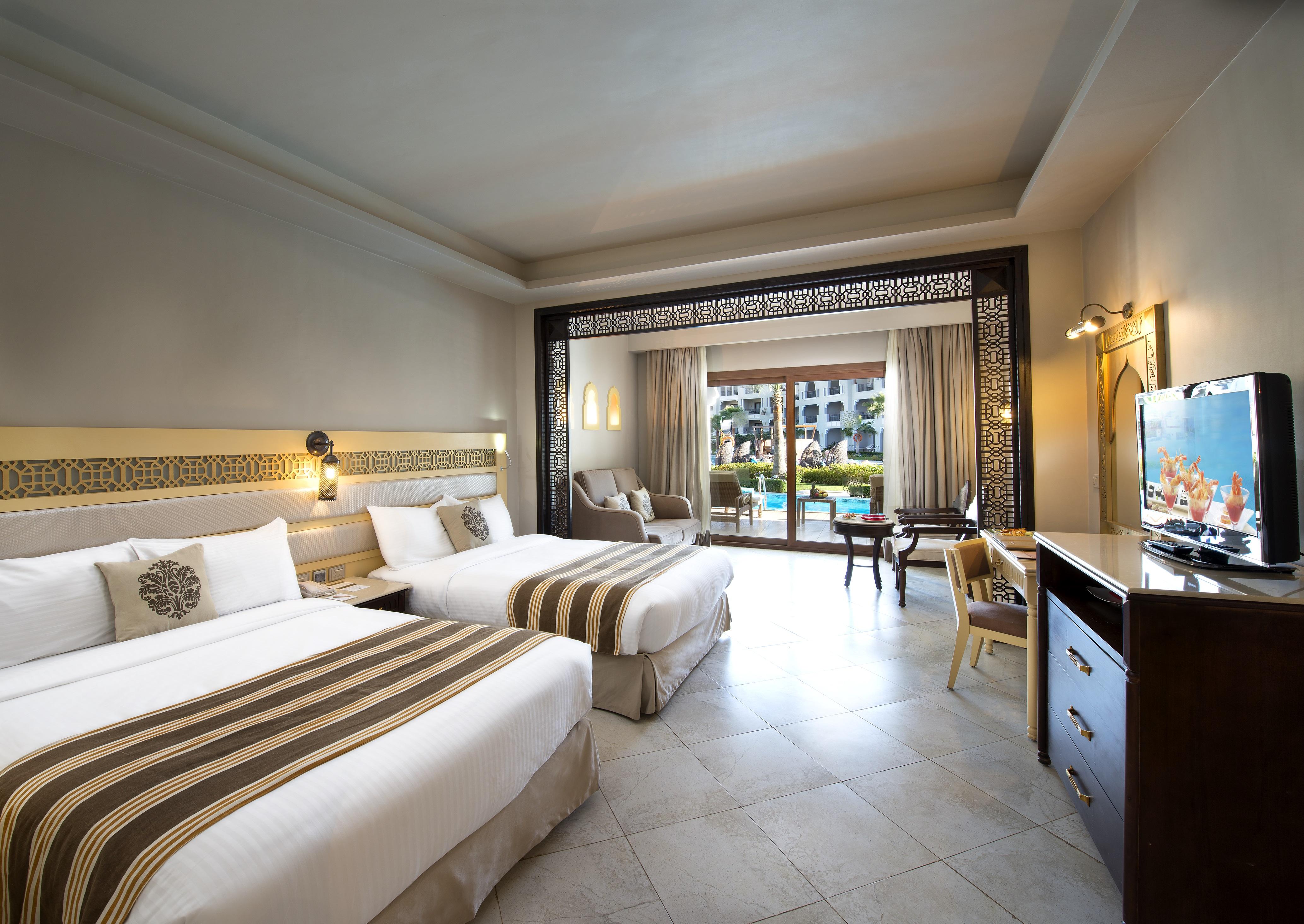 Sunrise Arabian Beach Resort Sharm el Sheikh Esterno foto The photo shows a well-decorated hotel room. It features two beds with white linens and decorative brown striped accents. There is a large window with sheer curtains that provides a view of the outdoors, suggesting a pool area or gardens. The room in