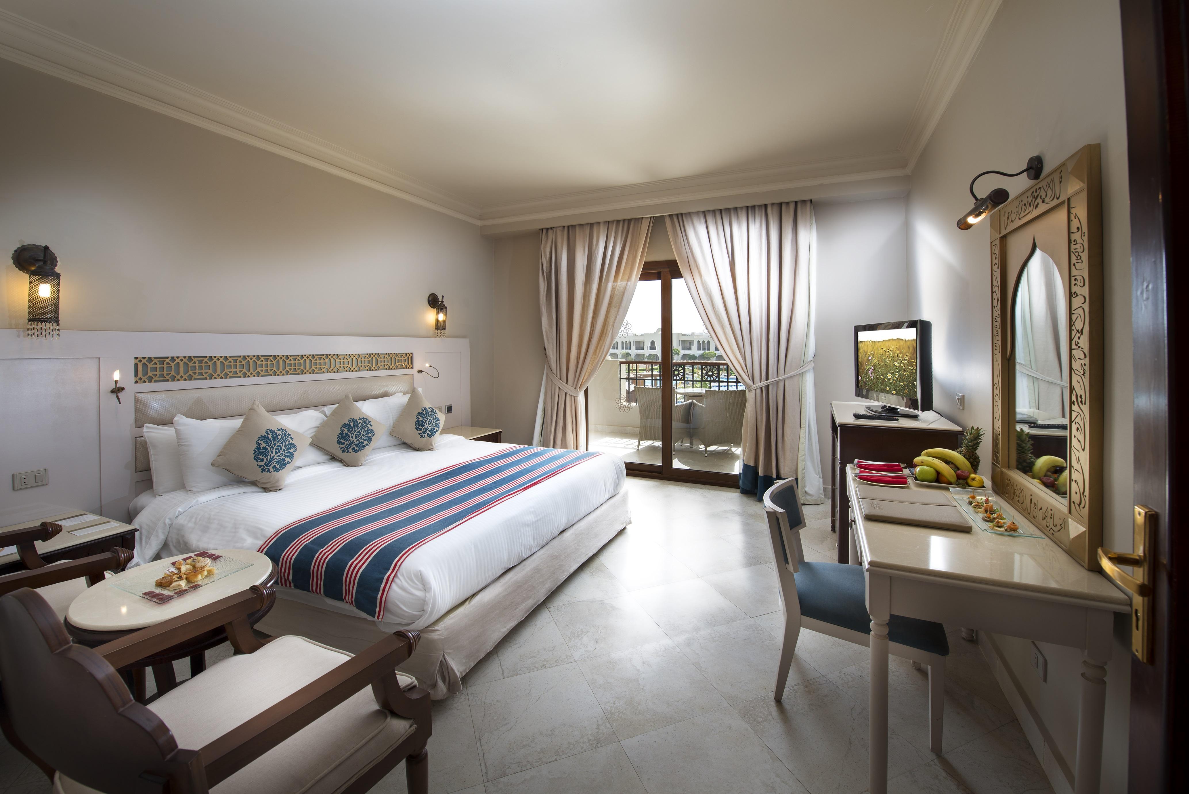 Sunrise Arabian Beach Resort Sharm el Sheikh Esterno foto The photo depicts a hotel room that appears stylish and well-furnished. It features a large bed with decorative pillows and a colorful blanket. There is a small seating area with armchairs and a round table, as well as a work desk with a chair. A tel
