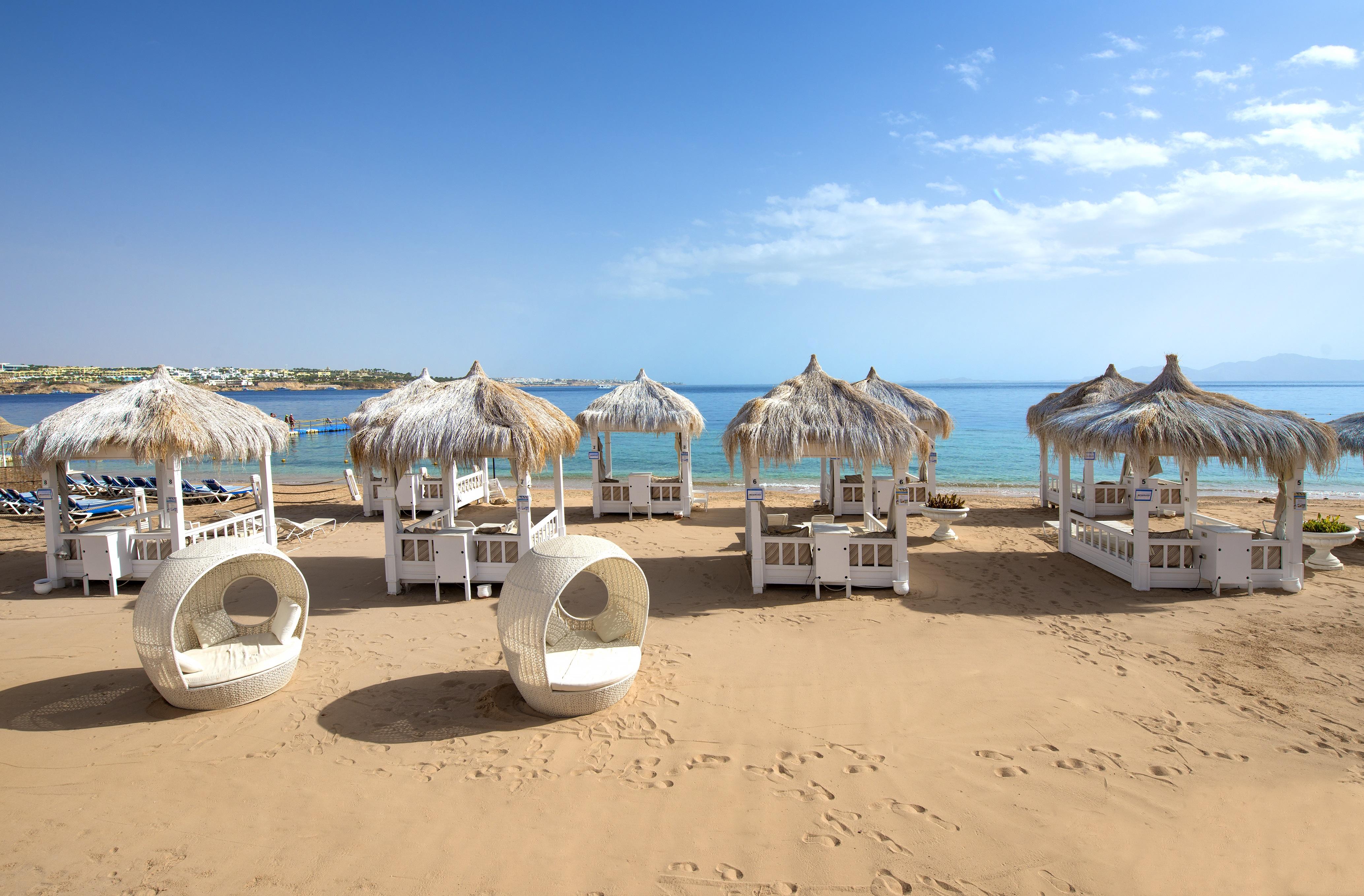 Sunrise Arabian Beach Resort Sharm el Sheikh Esterno foto The photo shows a picturesque beach scene featuring several stylish white cabanas with thatched roofs, set on golden sand. They are arranged neatly, with some circular lounge chairs nearby. In the background, the calm blue sea stretches out under a c