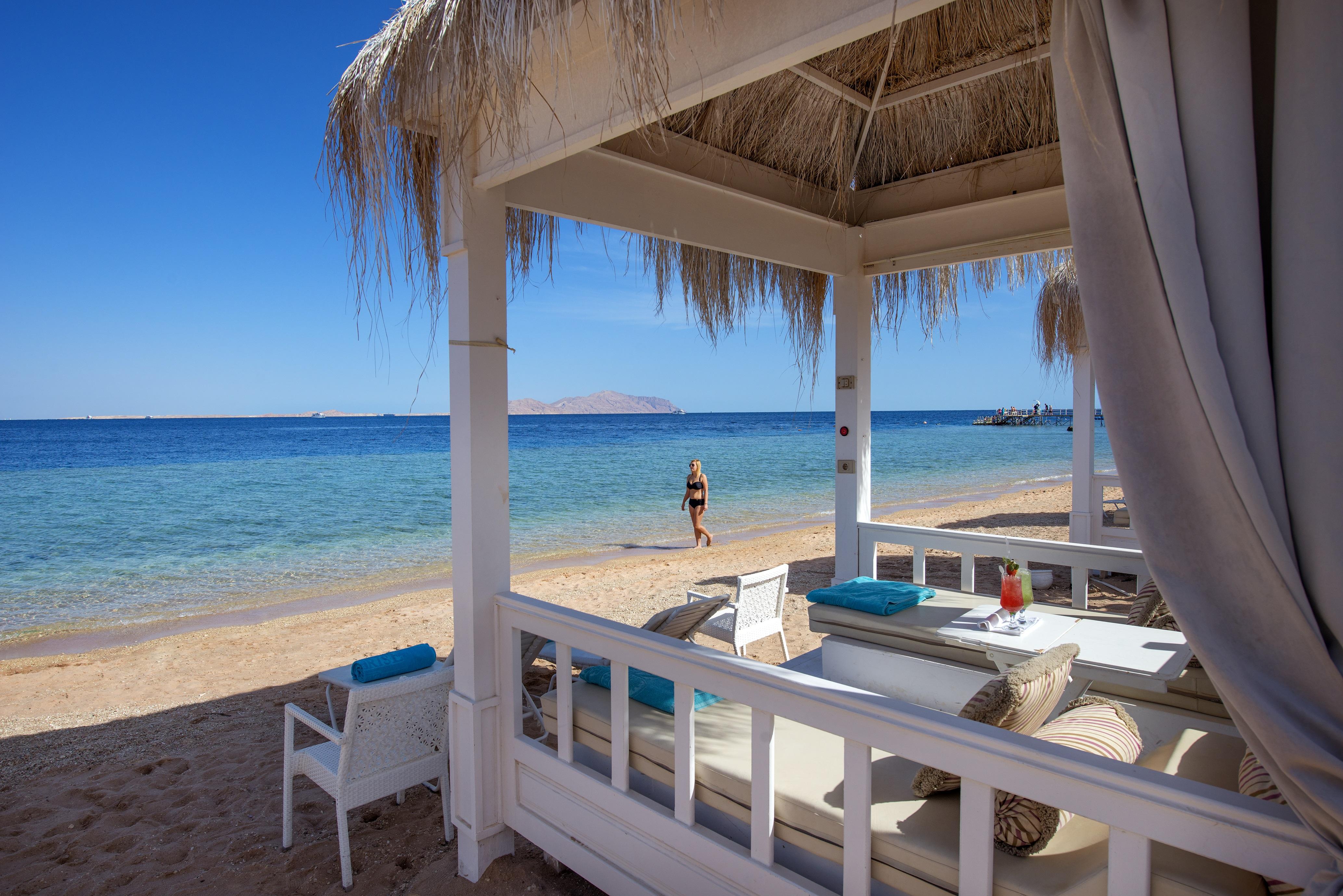 Sunrise Arabian Beach Resort Sharm el Sheikh Esterno foto The photo depicts a serene beach scene. In the foreground, there is a shaded seating area with white furniture and blue accents, creating a relaxing atmosphere. Behind the seating area, a sandy beach leads to calm, clear blue waters. A person is walk