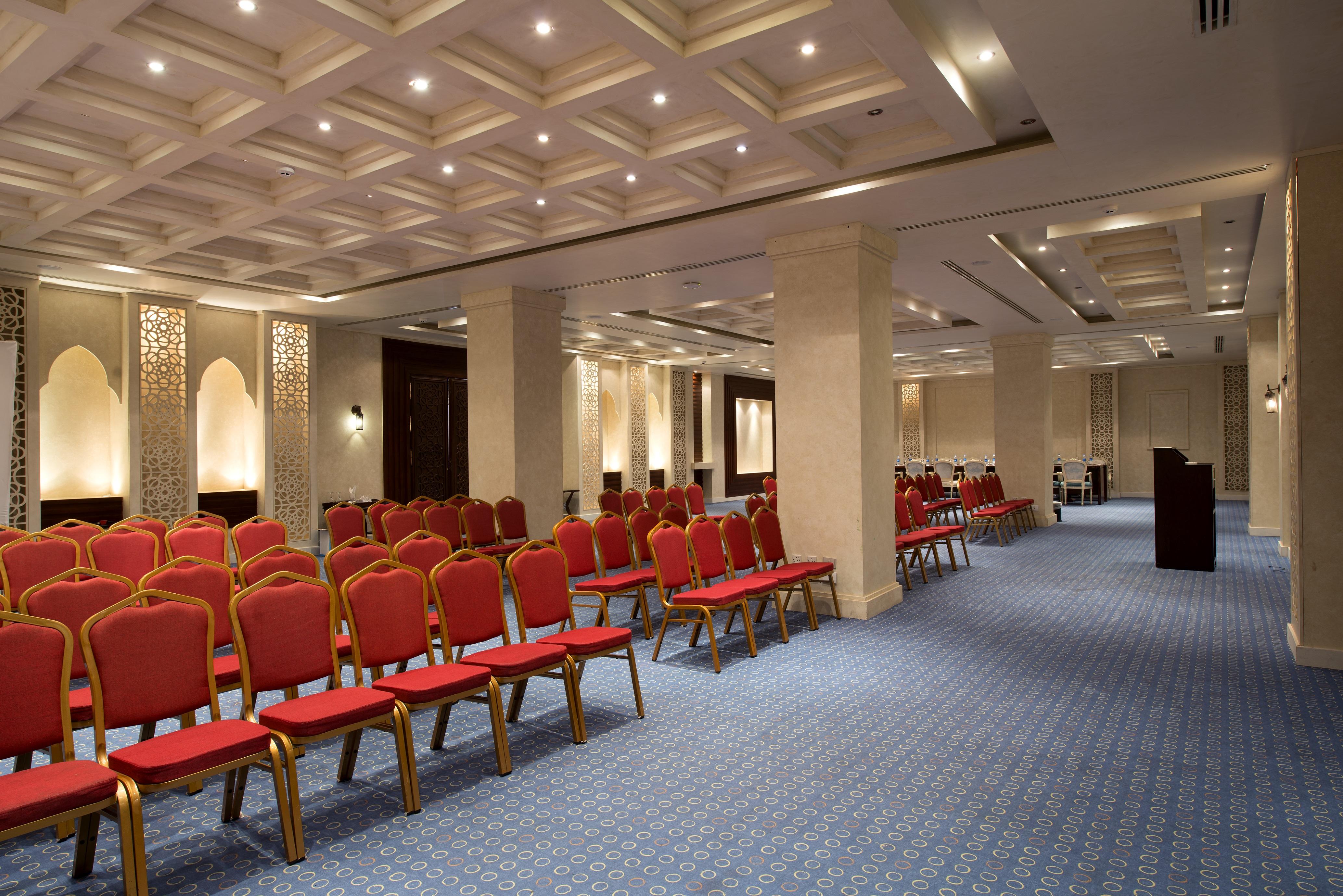 Sunrise Arabian Beach Resort Sharm el Sheikh Esterno foto The image shows a spacious indoor venue designed for meetings or events. It features rows of red chairs arranged neatly on a blue carpeted floor. The walls are adorned with decorative panels, and the ceiling has a modern geometric design with recesse