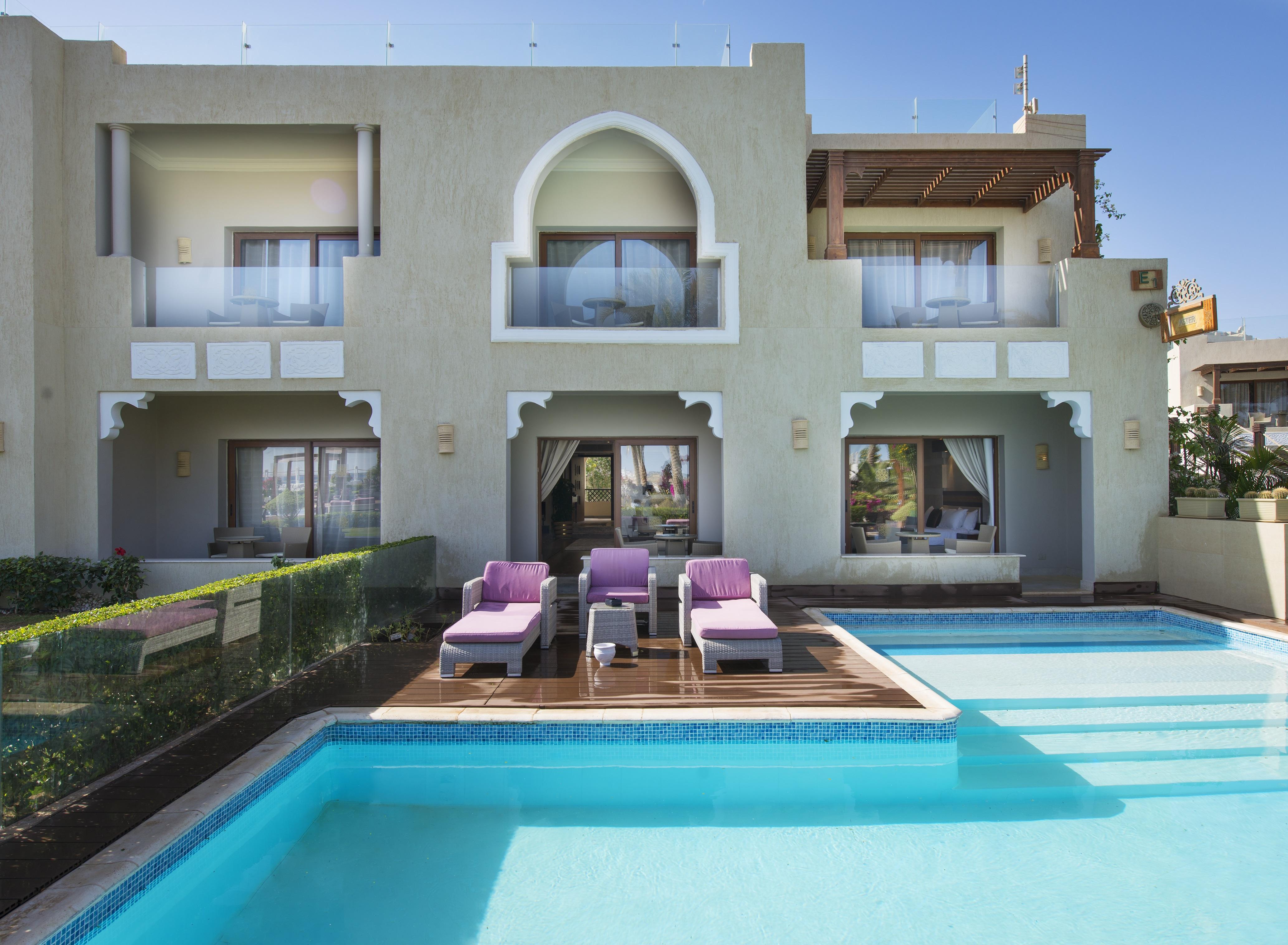 Sunrise Arabian Beach Resort Sharm el Sheikh Esterno foto The photo shows a luxurious modern villa with an elegant architectural style. In the foreground, there is a swimming pool that is clear and inviting, surrounded by a wooden deck. There are several lounge chairs with purple cushions positioned near th