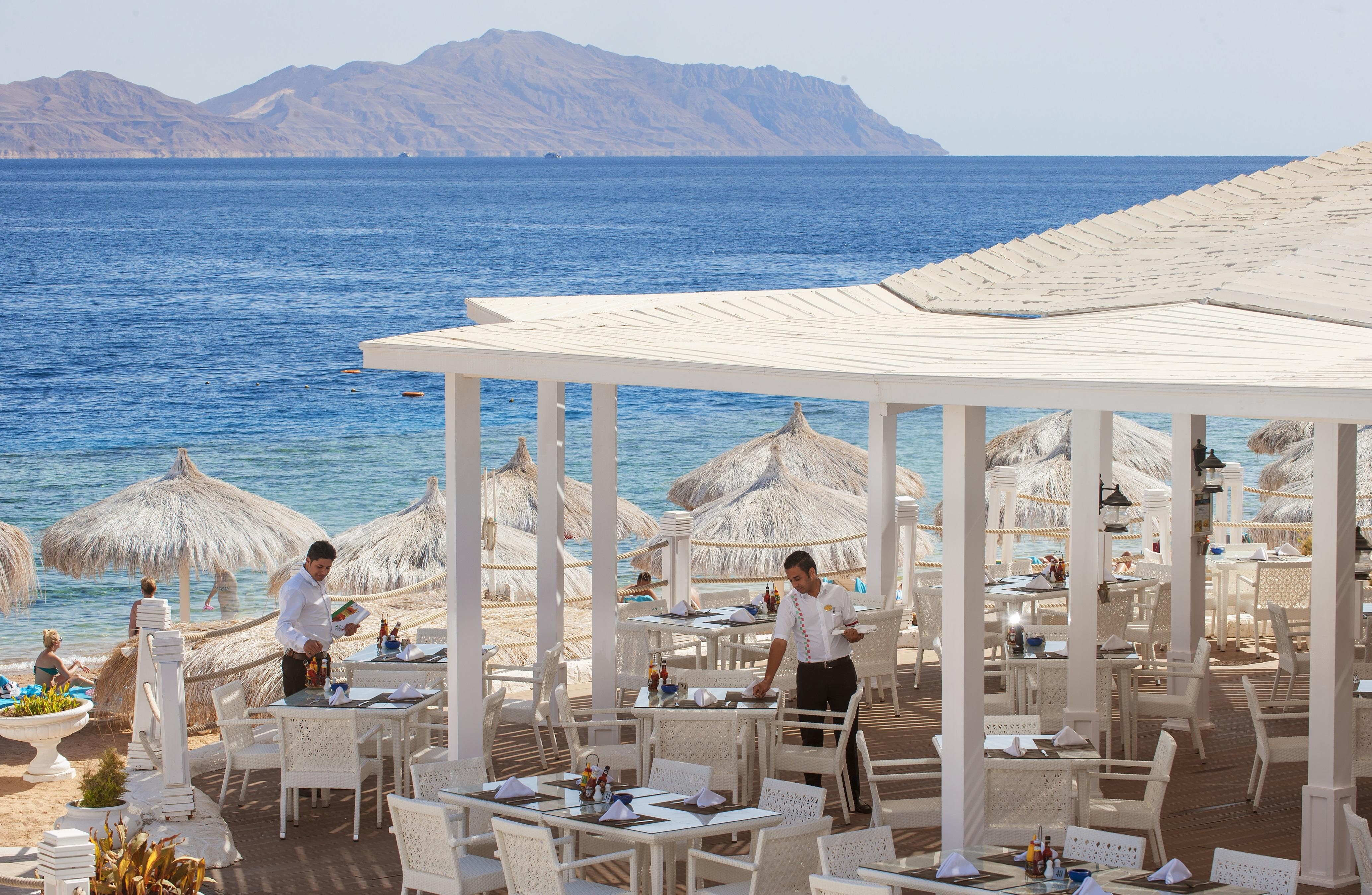 Sunrise Arabian Beach Resort Sharm el Sheikh Esterno foto The photo depicts a seaside restaurant with a beautiful view of the ocean and distant mountains. The outdoor dining area features white tables and chairs arranged under straw umbrellas for shade. Two waitstaff members are visible, attending to the ta