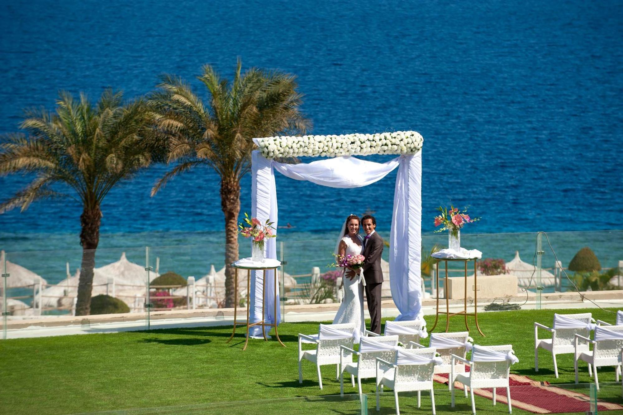 Sunrise Arabian Beach Resort Sharm el Sheikh Esterno foto The photo depicts a wedding scene set against a beautiful seaside backdrop. In the foreground, a couple is standing underneath a decorative wedding arch adorned with white fabric and flowers. The groom is in a dark suit, while the bride is dressed in