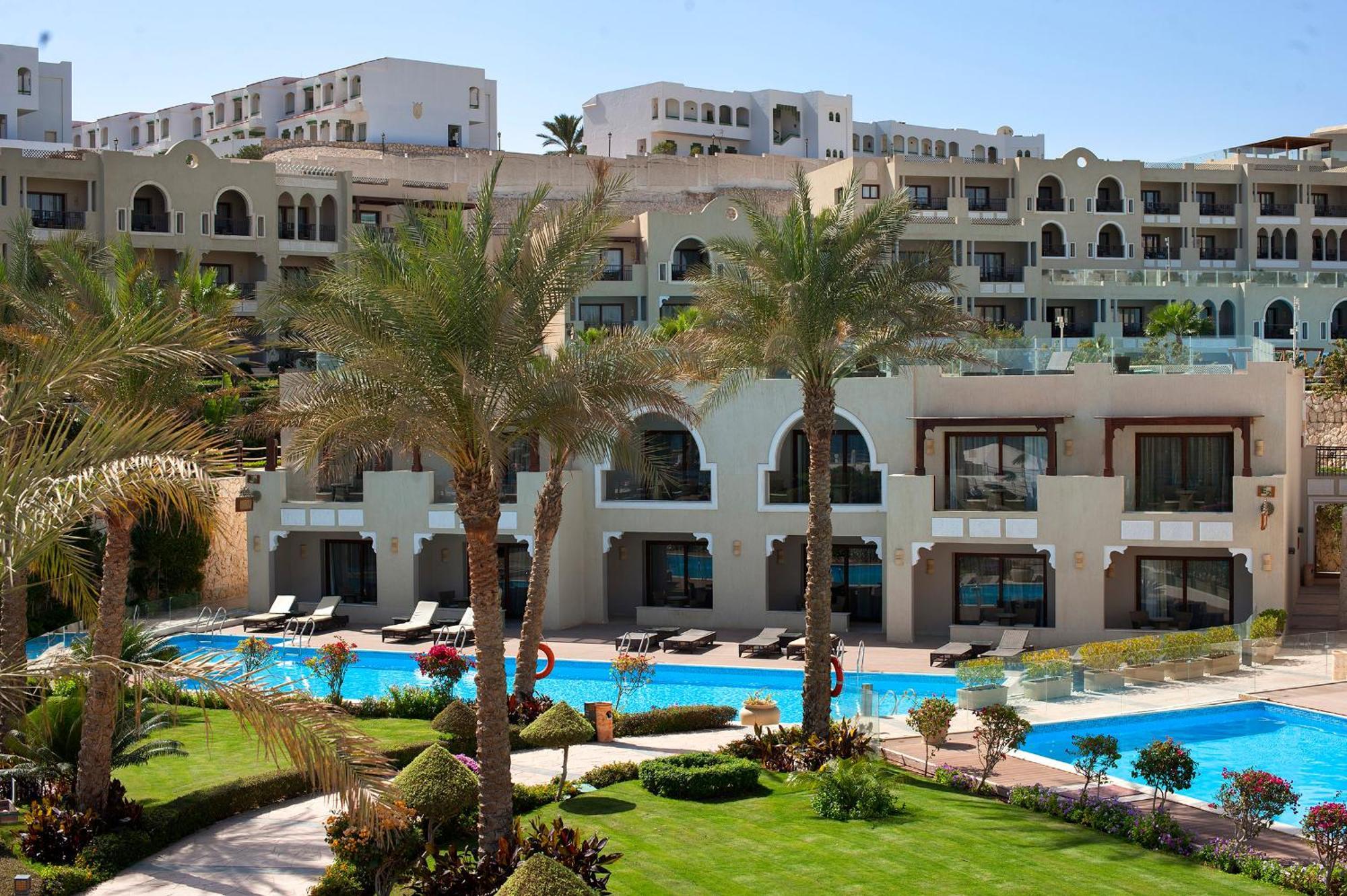 Sunrise Arabian Beach Resort Sharm el Sheikh Esterno foto The photo depicts a luxurious resort or hotel complex. In the foreground, there is a landscaped area with manicured lawns and palm trees. A swimming pool is visible, reflecting the clear blue sky. The background features several multi-story buildings