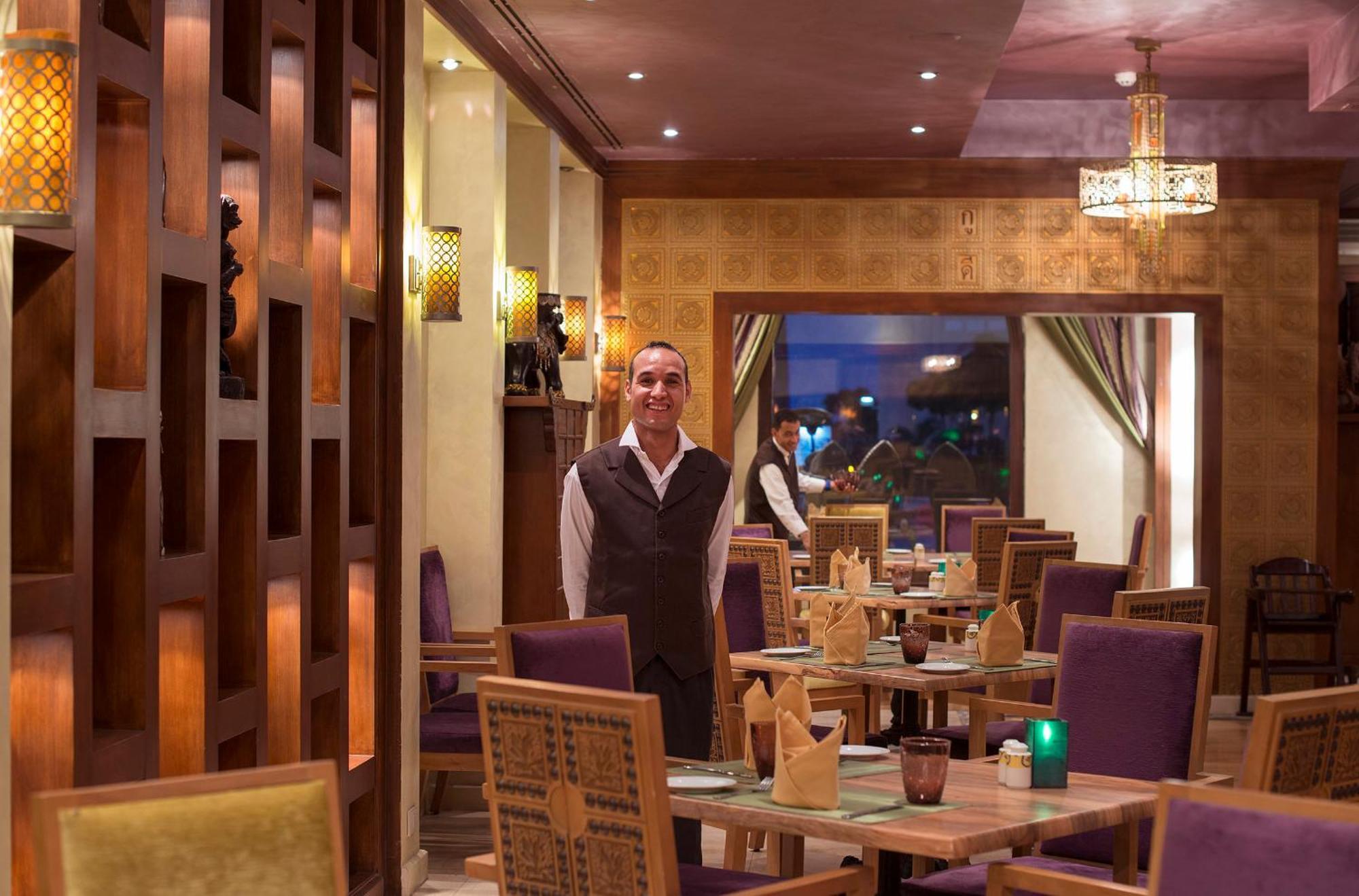 Sunrise Arabian Beach Resort Sharm el Sheikh Esterno foto The photo depicts the interior of a restaurant featuring a staff member standing in the foreground. The restaurant has a warm and inviting ambiance, characterized by wooden accents, stylish lighting, and neatly arranged tables. The seating includes p